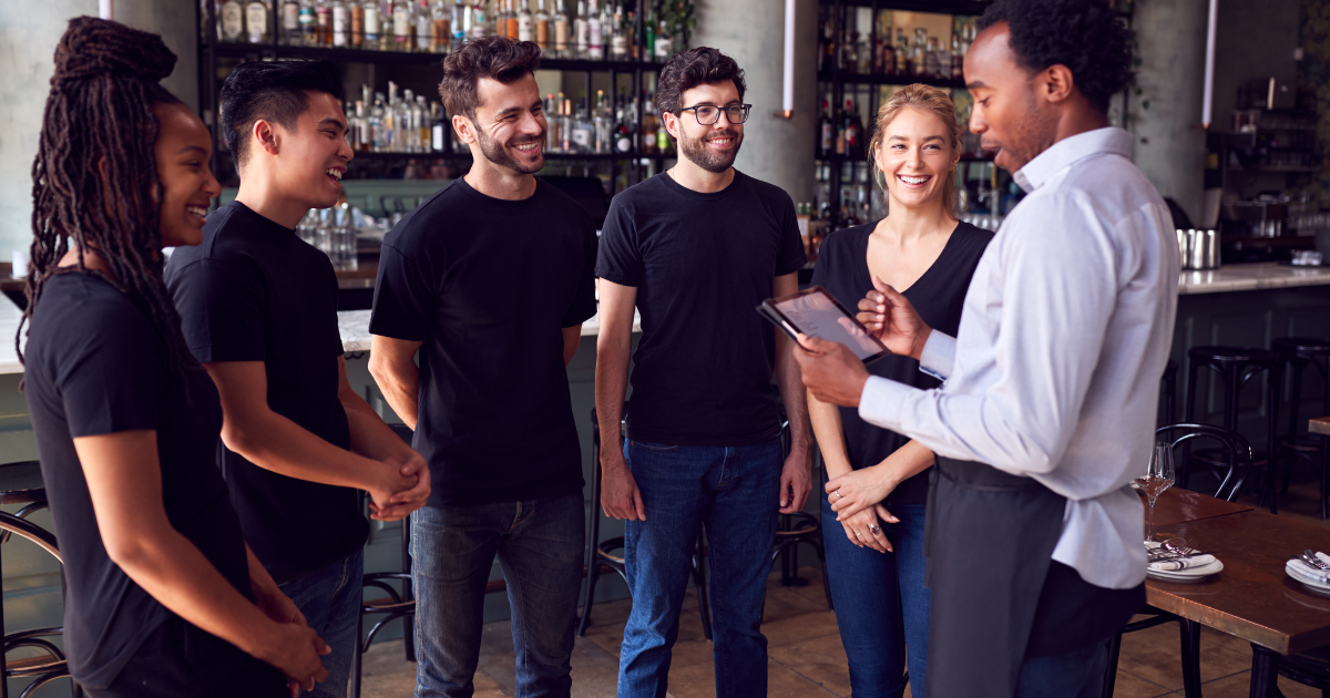 Imagem contendo seis funcionários de um bar, sendo uma mulher negra, três homens brancos, uma mulher branca e um homem negro. O homem negro aparenta ser o gerente e passa informações aos demais. Todos estão sorridentes.